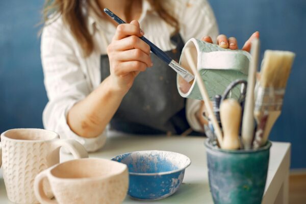 personnage féminin sur fond bleu, assise tenant un pinceau et une tasse qu'elle décore. Bout de table beige avec mug beige et bleu ainsi que plusieurs panneaux